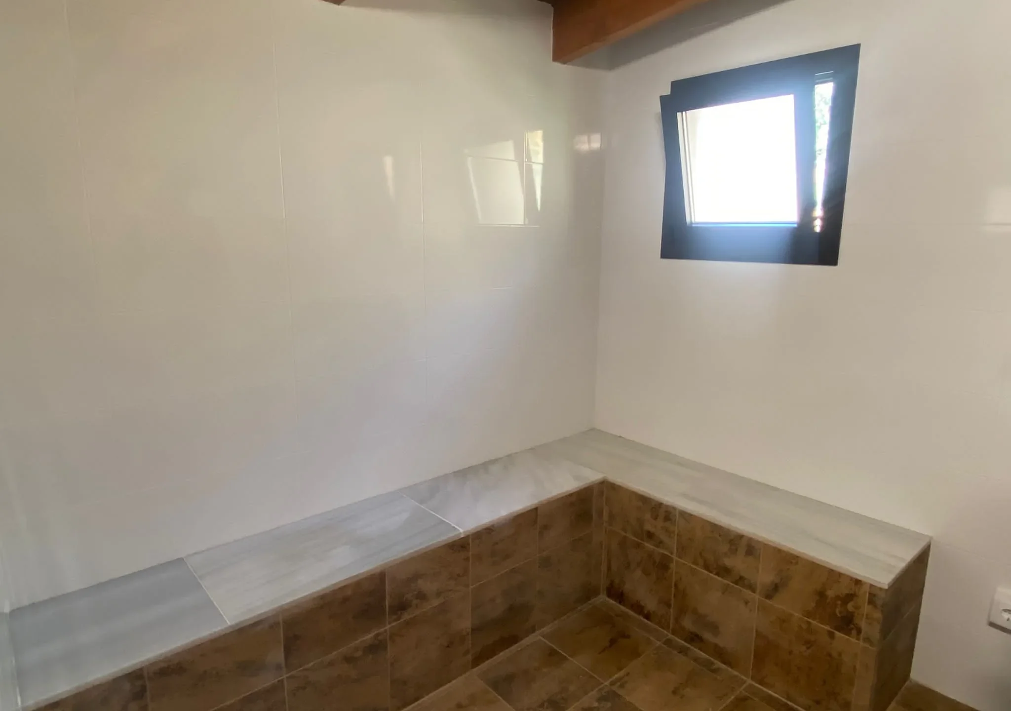 Baño de vapor con banco corrido en L de piedra clara, paredes de azulejo blanco y ventana