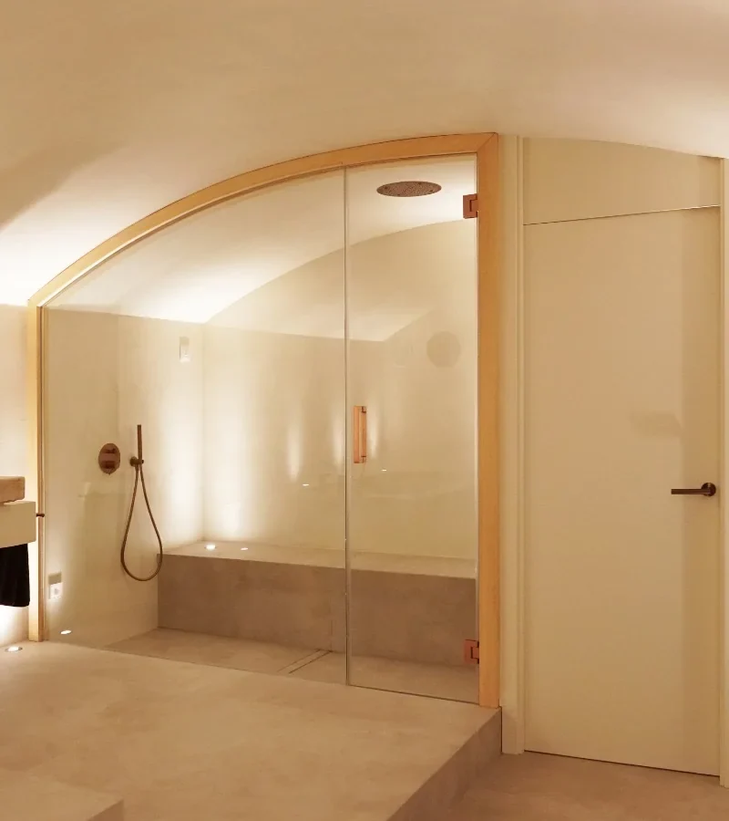 Baño de vapor de microcemento tras una mampara de cristal, con banco corrido, rociador de techo y un lavabo de piedra a la izquierda.