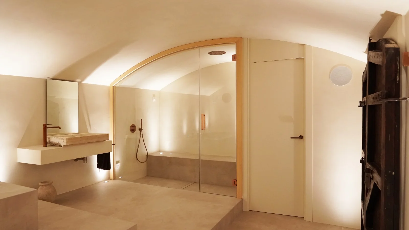 Baño de vapor de microcemento tras una mampara de cristal, con banco corrido, rociador de techo y un lavabo de piedra a la izquierda.