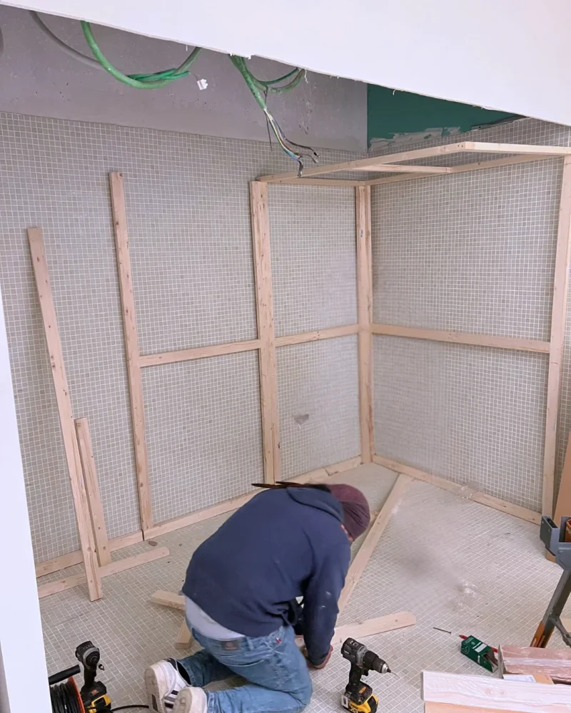 Un carpintero, de rodillas en el suelo, atornilla los travesaños del bastidor de una sauna contra la pared alicatada; alrededor, atornilladores y listones aún sin colocar.