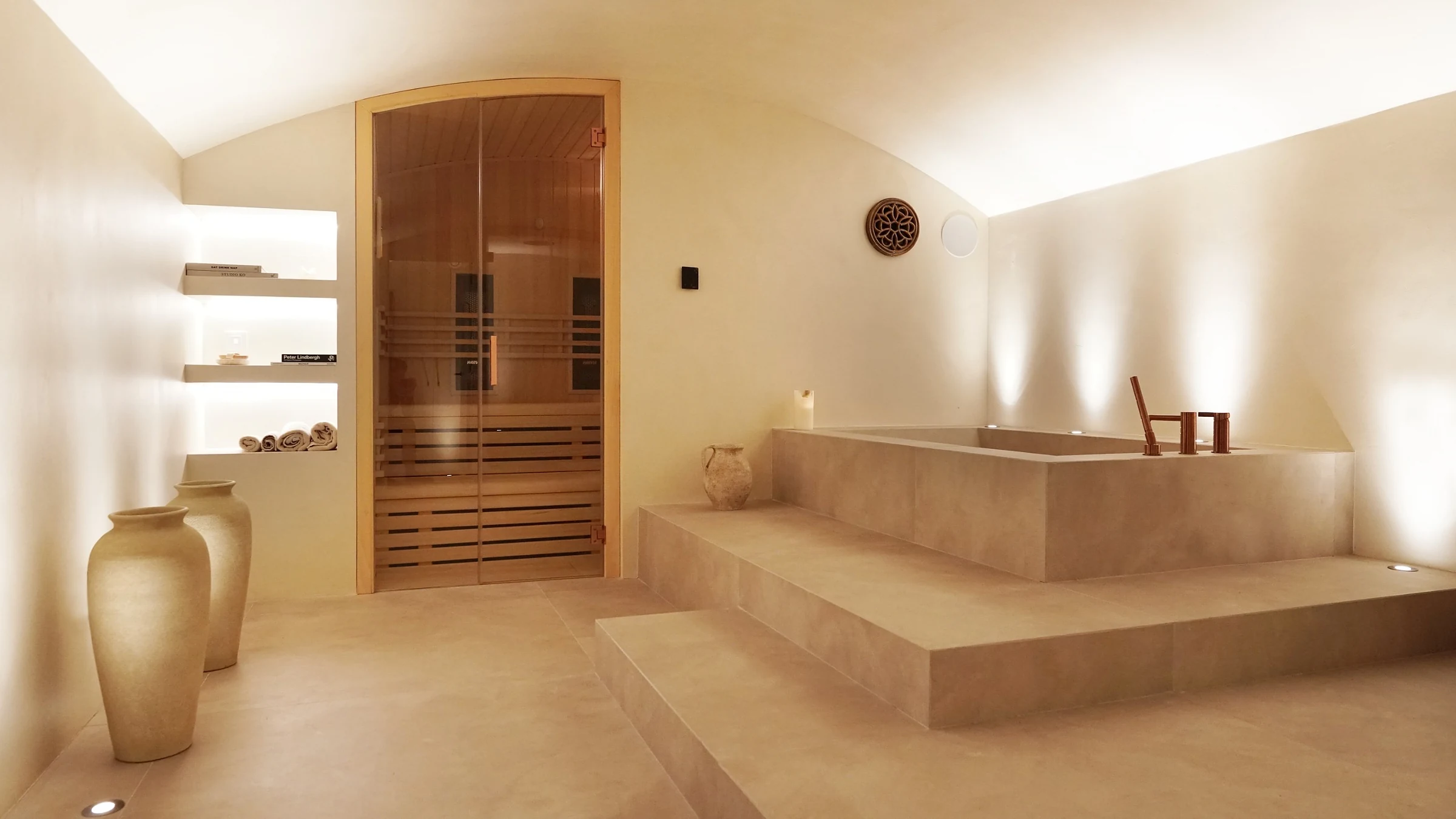 Sala de hidroterapia con bóveda de microcemento: al fondo, una cabina de sauna con puerta de cristal; a la derecha, una bañera de obra encastrada entre dos escalones.