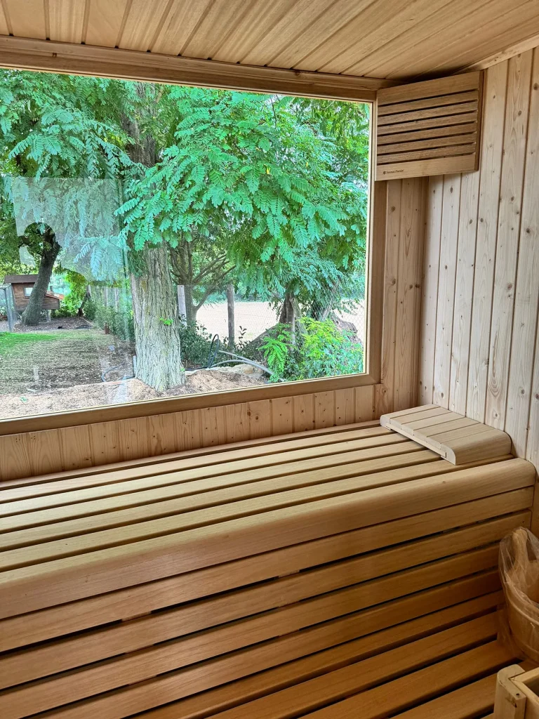 Banco corrido de una sauna de exterior con reposacabezas de listones y, tras el ventanal, el jardín arbolado.