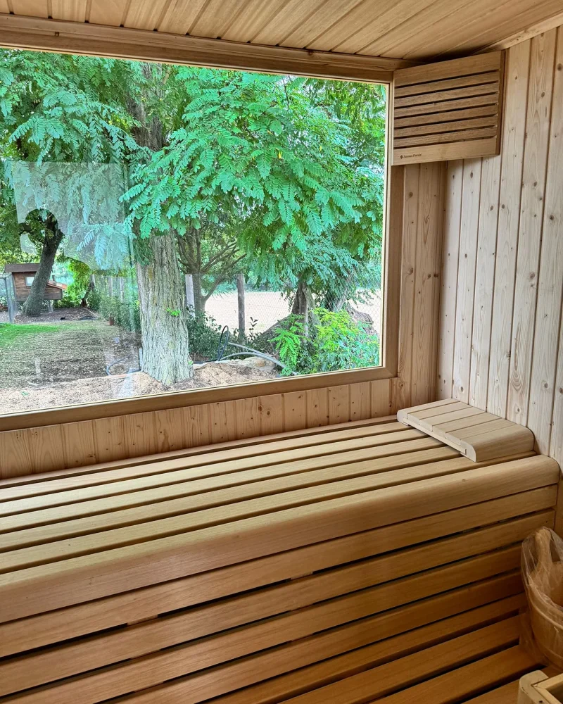 Banco corrido de una sauna de exterior con reposacabezas de listones y, tras el ventanal, el jardín arbolado.