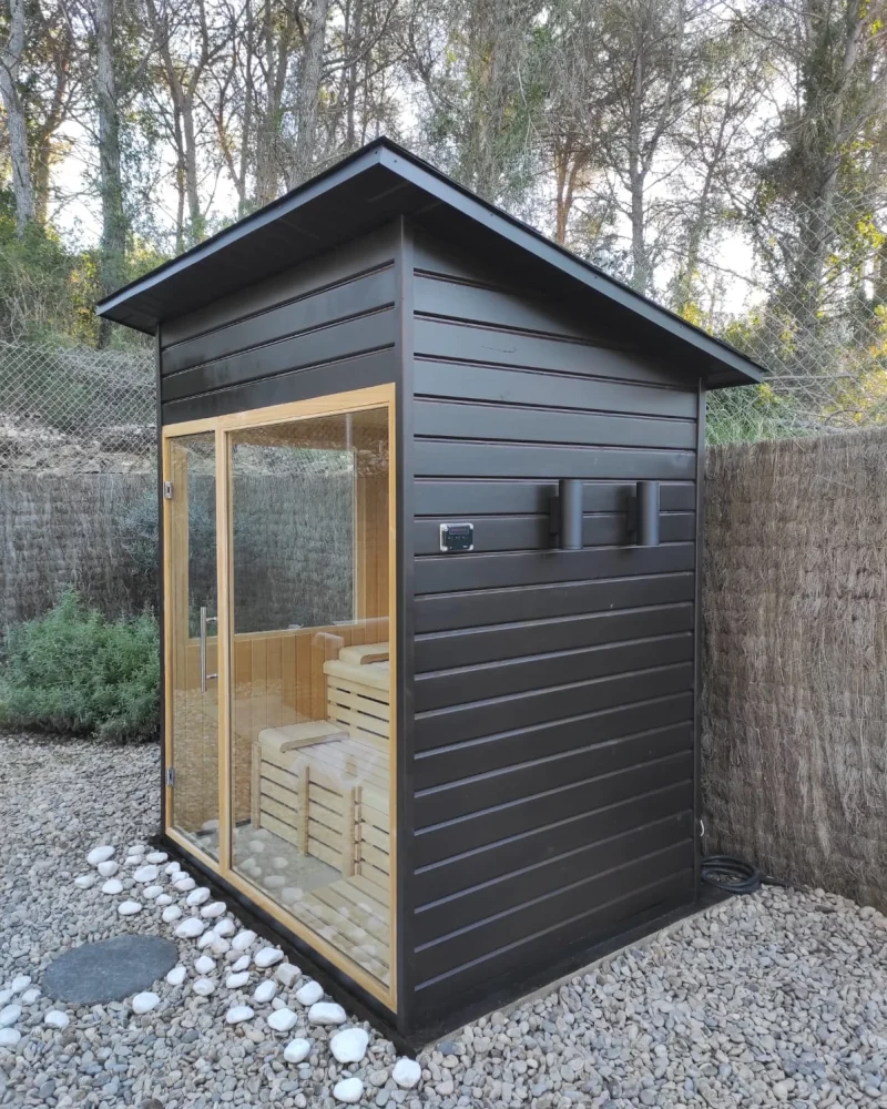 Sauna de exterior con el revestimiento pintado de negro y un frente acristalado que deja ver los bancos de madera clara, sobre un lecho de grava.