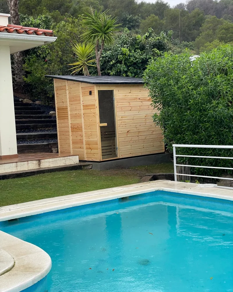 Caseta de sauna de exterior en madera clara con cubierta plana, levantada sobre el césped junto a una piscina.
