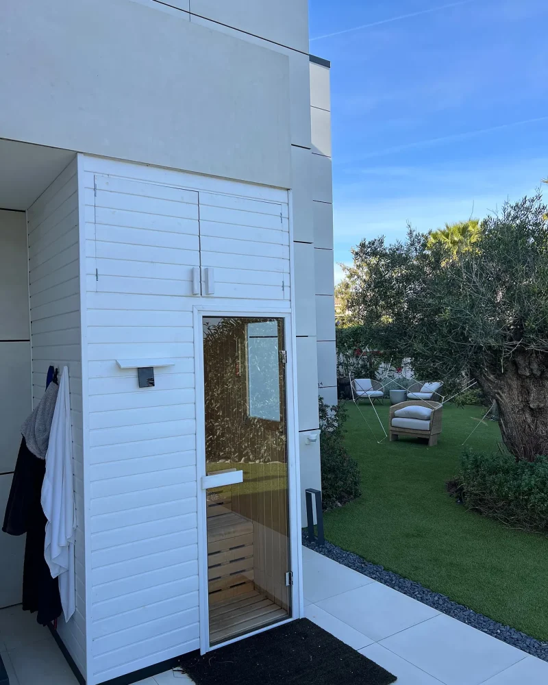 Módulo de sauna revestido de madera lacada en blanco y puerta de cristal, adosado a la fachada de una casa con césped y un olivo.