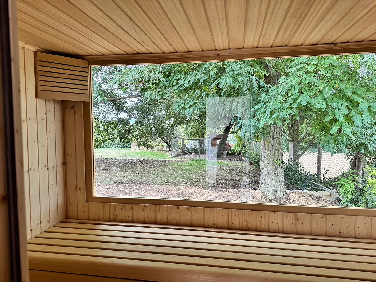 Interior de una sauna de exterior visto desde el banco: un ventanal corrido ocupa toda la pared y deja ver el jardín y el tronco de una acacia.