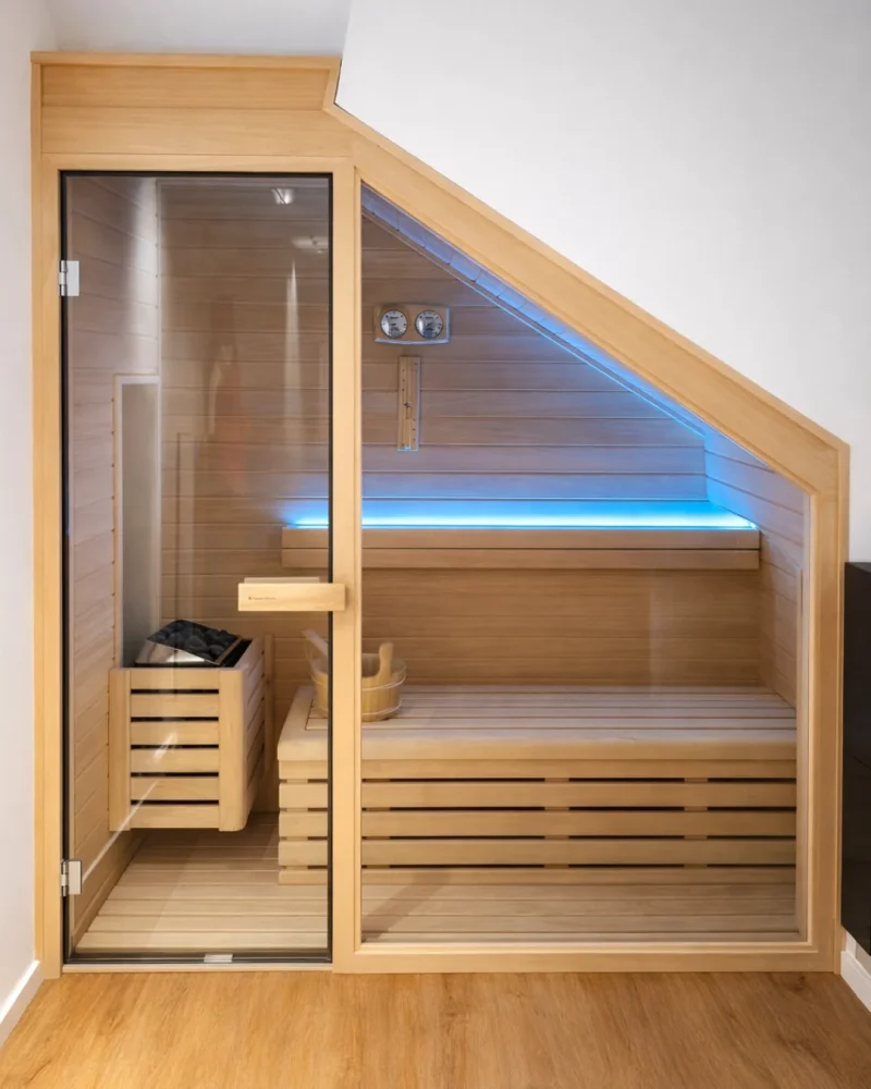 Sauna construida bajo el tiro de una escalera: el banco superior sigue la diagonal del peldañeado y una tira de luz azul marca el corte.