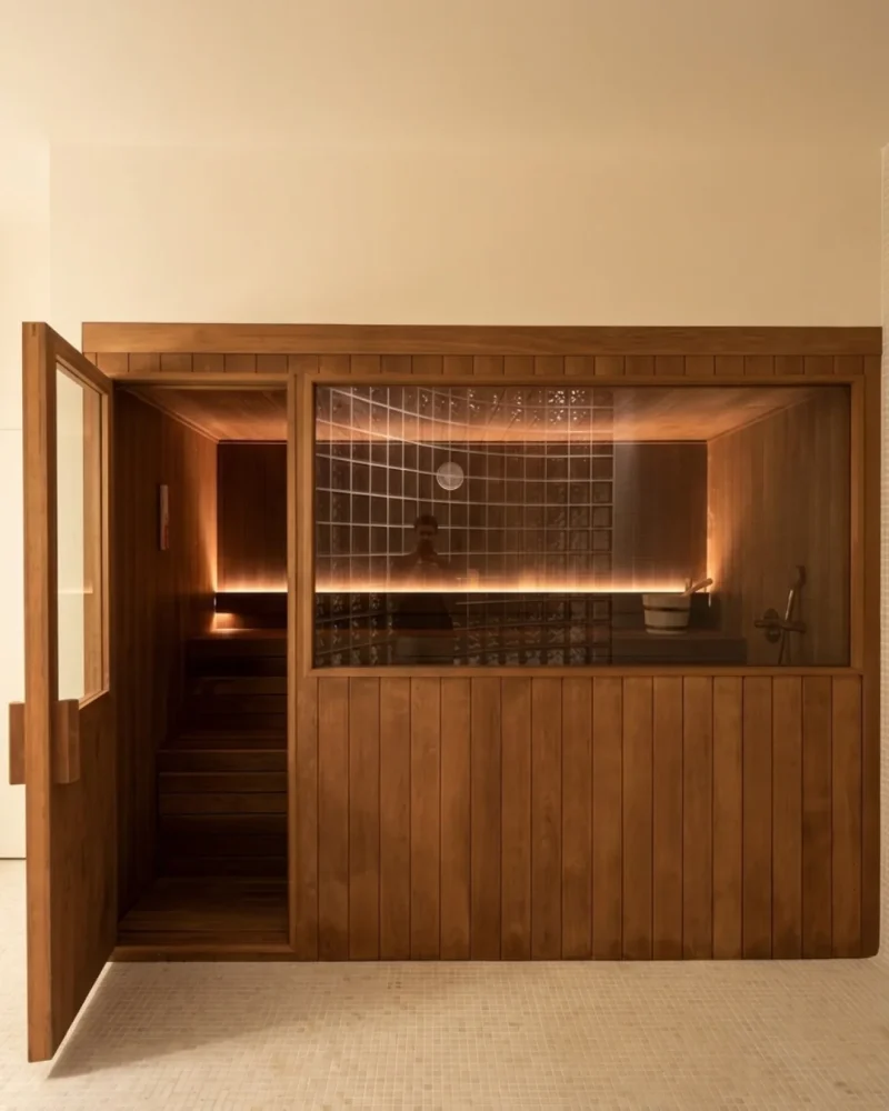 Cabina de sauna en madera termotratada oscura con un gran cristal fijo, montada contra una pared de pavés.