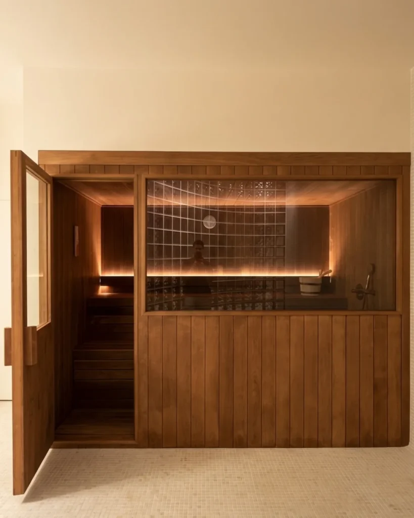 Cabina de sauna en madera termotratada oscura con un gran cristal fijo, montada contra una pared de pavés.