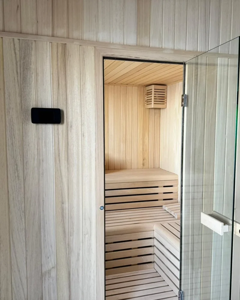 Frente de una sauna de interior en madera clara con la puerta de cristal abierta y el mando de control empotrado en el revestimiento.
