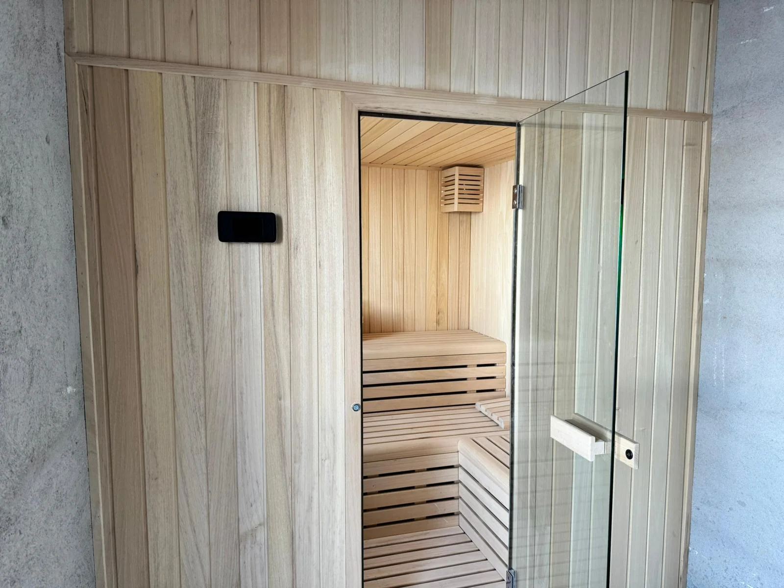 Frente de una sauna de interior en madera clara con la puerta de cristal abierta y el mando de control empotrado en el revestimiento.