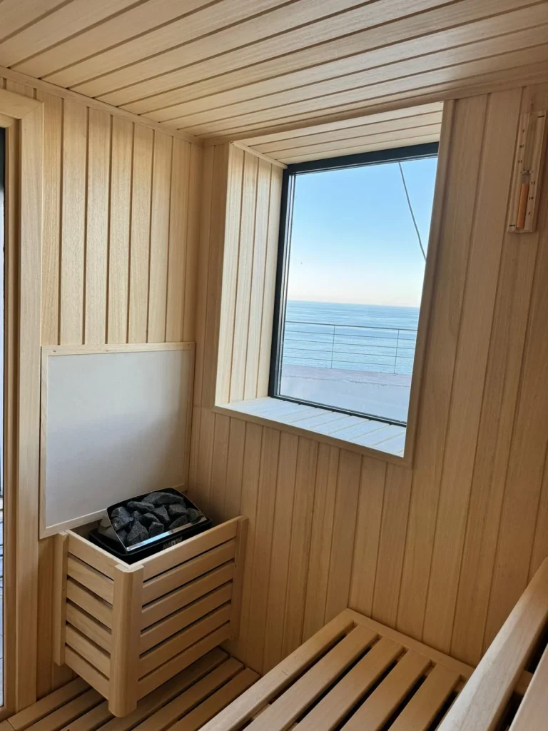 Rincón de una sauna con la estufa de piedras junto al banco y una ventana cuadrada abierta al mar.