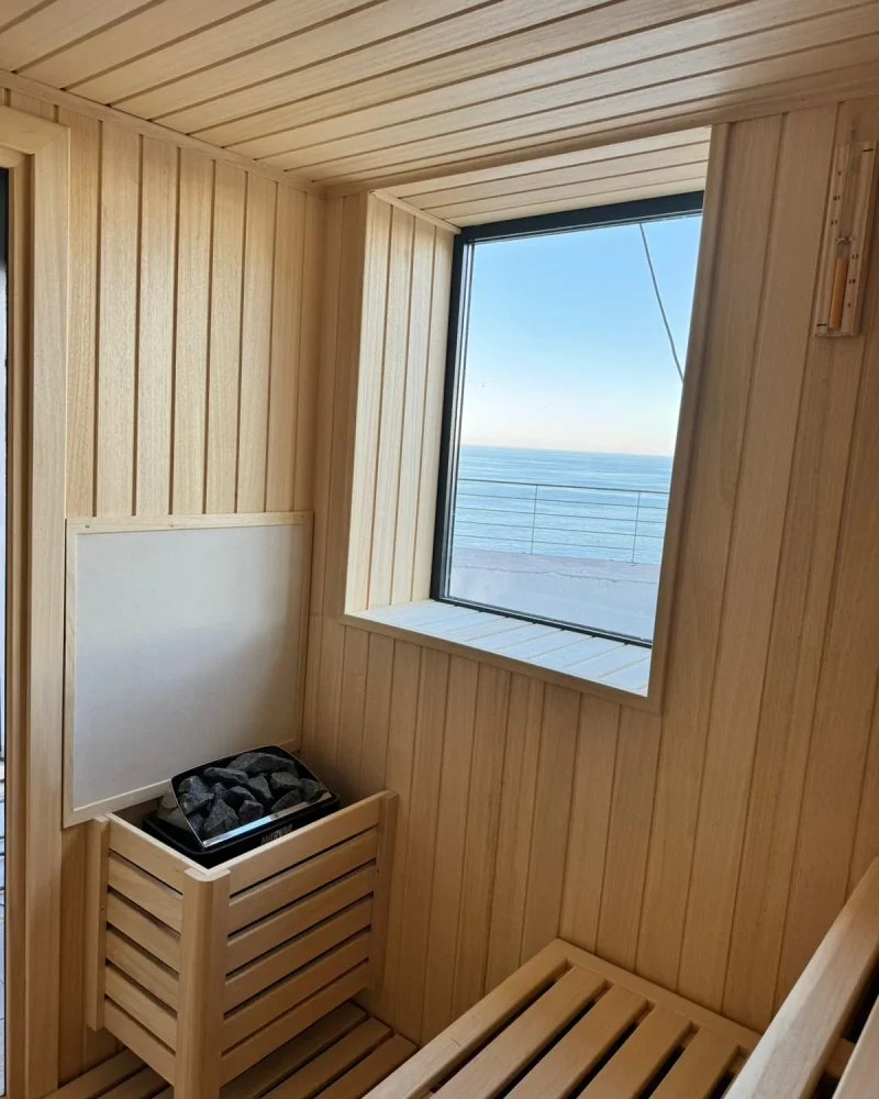 Rincón de una sauna con la estufa de piedras junto al banco y una ventana cuadrada abierta al mar.